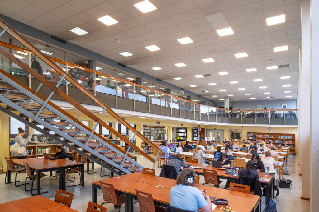 Interior of a modernist library reading room, a rectangular room filled with tables on which patrons are working. The room is surrounded by a mezzanine reached by a metallic crossing into the room.