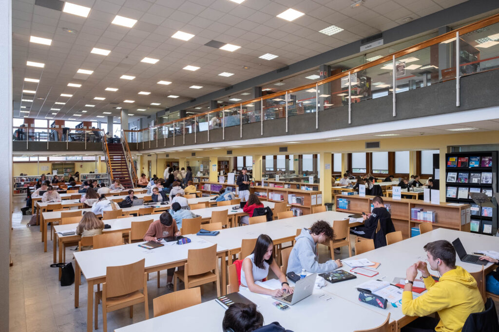 Interior of a modernist library reading room, a rectangular room filled with tables on which patrons are working. At the back of the room is an angled stair reaching to a wraparound mezzanine.