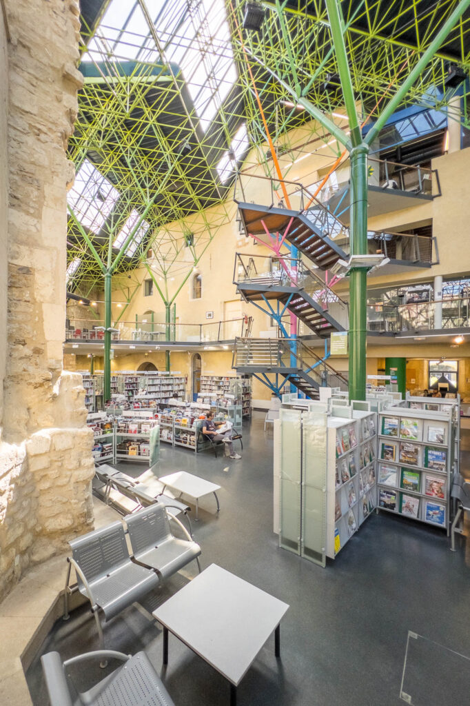 Interior of a library inside a converted former hospital, a high glass roof on metal stilts covers an area bound by original stone walls. The metal stilts branch out as they rise and are painted green, recalling tree trunks.