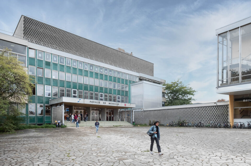 Exterior of a Modernist university campus showing a four-story rectangular building with a facade of alternating bands of windows and green metal panels. The building is topped with a two-story structure covered with a concrete openwork screen. The same screen is repeated in front of a low building to the right of the main structure, and on either side of its porte cochère, on which the words "Faculté de médecine" can be read. The word "Bibliothèque" is stencilled one letter at a time on the top windows. Students are show exiting the building on a paved plaza in front.