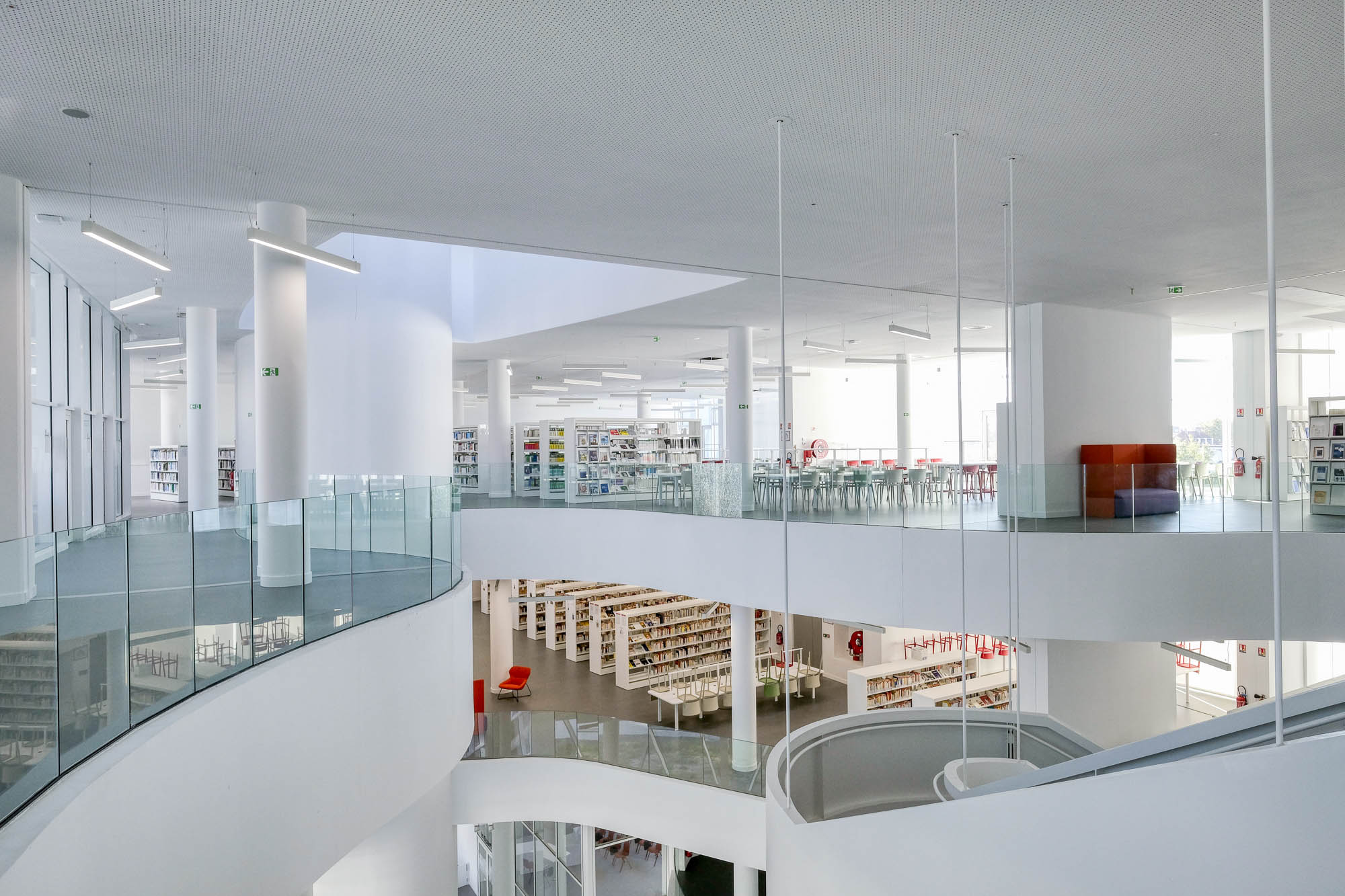 A contemporary, multilevel library with curved balconies, ramps and stairs. Filled bookshelves and colourful seats contrast against the grey and white surroundings.