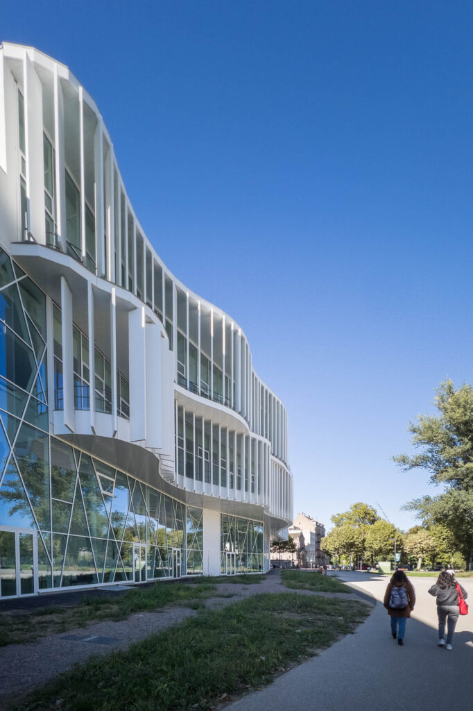 Exterior of a contemporary library, a white curvy building with vertical brise-soleils. People are walking on a path in the front.