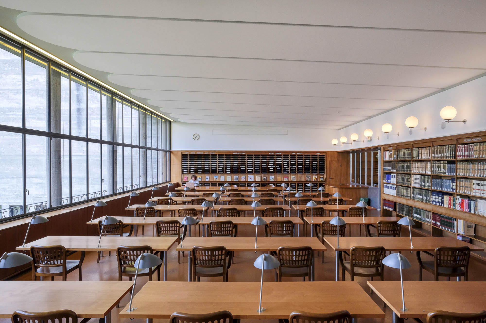 Interior of a modernist library reading room with a large window to the left. To the right is a long bookcase, with globe lights above it. The room is filled with tables topped with work lights. At the back is a magazine display rack. A single person sits at a table at the back.