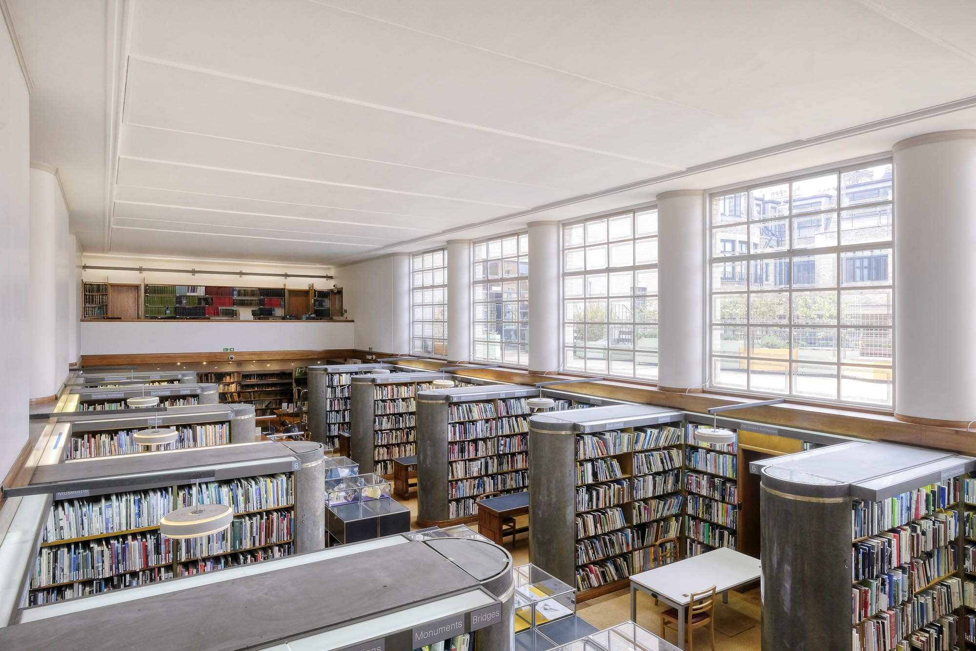 Interior of a library looking down from a mezzanine. The library has rows of bookshelves that end in a semicircular stone column. There are windows to the right, separated by half columns recalling those of the bookshelves.