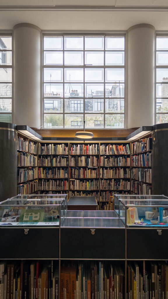 Interior of a library showing a book nook filled with bookshelves, with a row of windows above. In front are display cases with books underneath.