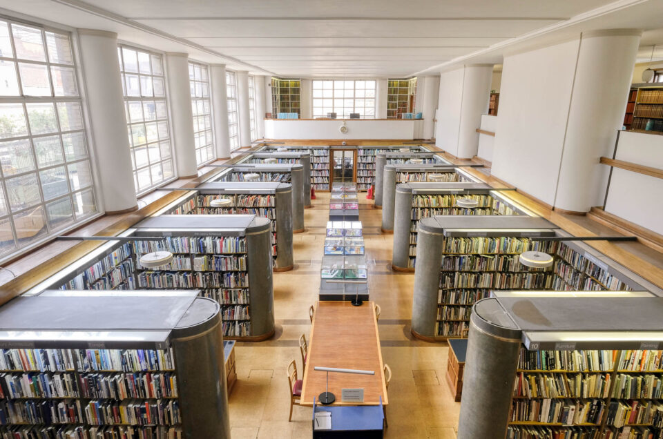 Interior of a library looking down from a mezzanine. The library has rows of bookshelves that end in a semicircular stone column. There are windows to the left, separated by half columns recalling those of the bookshelves.