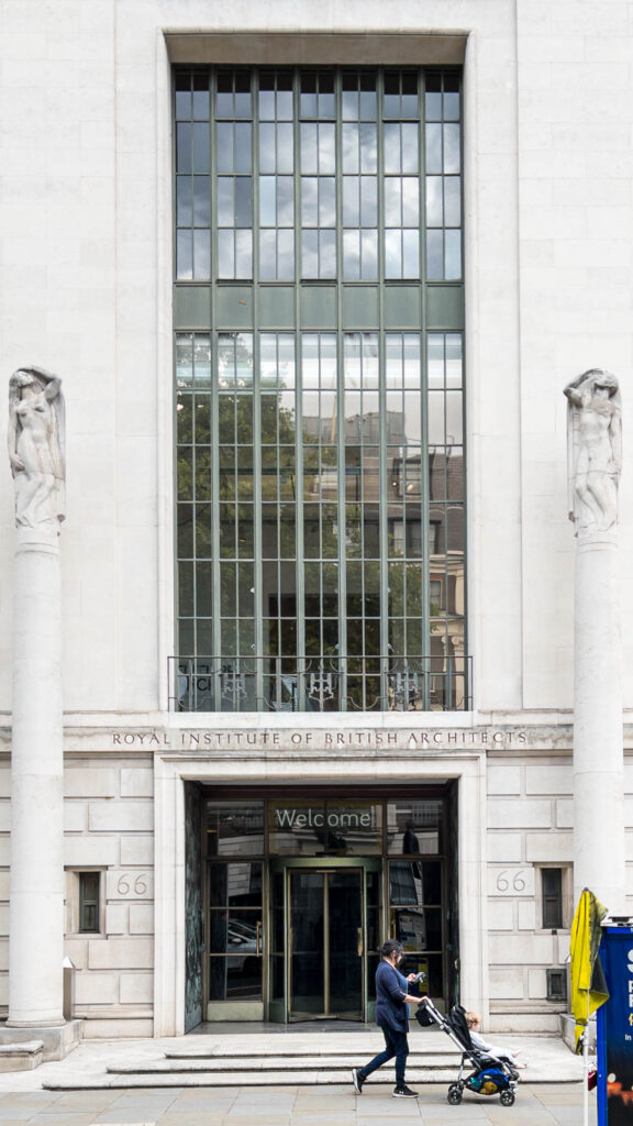 Exterior shot of an Art-Deco facade. The bottom third of the facade is rusticated and pierced by a square entrance, in which a rotating door is inset. The words "Welcome" are written above the door, and the number "66" is engraved on either sides of it. Above the entrance, the words "Royal Institute of British Architects" are engraved in a stone ledge. The above two-thirds of the facade are taken by a tall window. Two columns topped with sculptures of human figures stand on either sides of the entrance. A woman pushing a stroller is walking past the entrance.