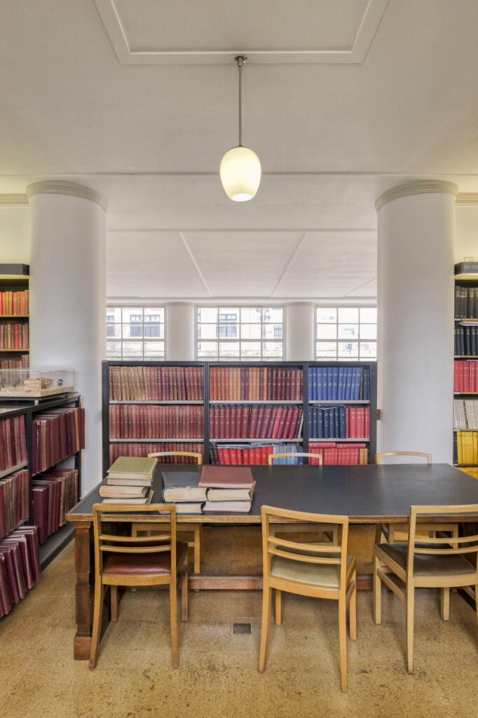 Interior of a library, with a table and six chairs, bookshelves and two columns.