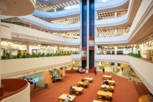 Interior of a multi-story library with open, tiered floors. Visible are several levels with bookshelves, study areas, and seating. A glass elevator is in motion in the background.