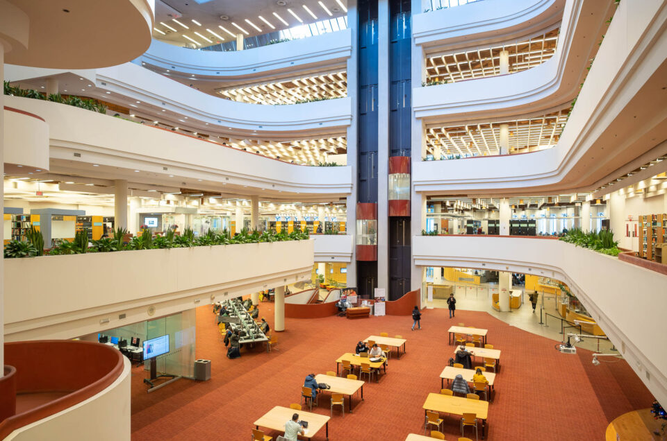 Interior of a multi-story library with open, tiered floors. Visible are several levels with bookshelves, study areas, and seating. A glass elevator is in motion in the background.