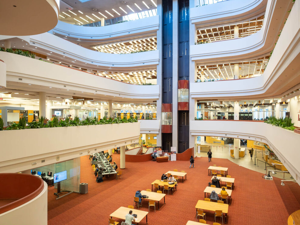 Interior of a multi-story library with open, tiered floors. Visible are several levels with bookshelves, study areas, and seating. A glass elevator is in motion in the background.