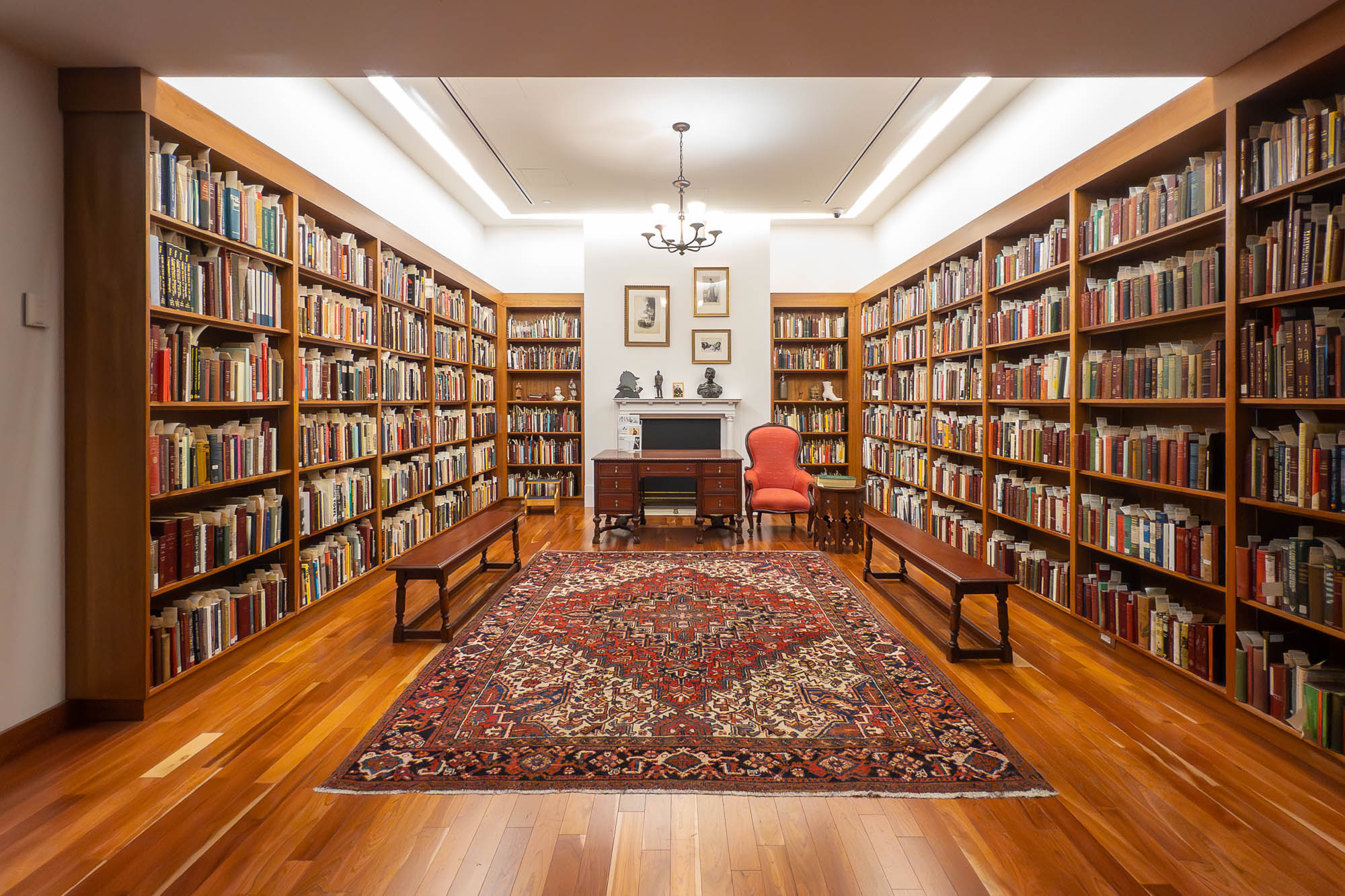 A library room with wooden shelves filled with books on both sides. A red patterned rug covers the wooden floor. At the far end, there is a small desk, an orange chair, and framed artwork on the wall above a mantelpiece.