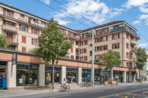 Exterior of an U-shaped 1920s multistory apartment building in Lausanne, Switzerland. Filling the courtyard in the middle of the building is a one-storey retail pavilion, now occupied by a public library. A cyclist is riding in front of the building.