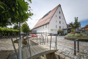 Exterior of a former granary transformed into a library. It is a steeply pitched four-storey whitewashed building with a red tiled roof. A small bridge over a paved brook is in the foreground.