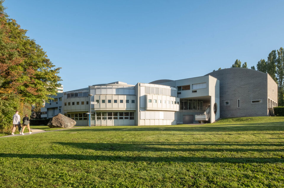 Exterior of a postmodern building, comprised of curved sections fanning out towards a grass meadow. Two people are walking on a path to the left.