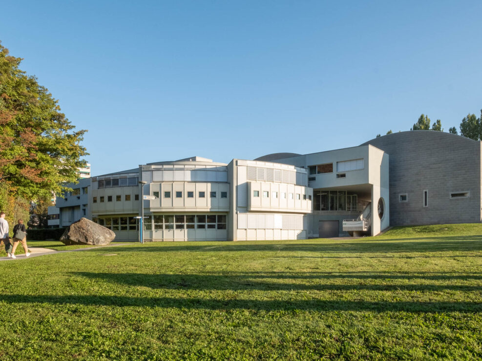 Exterior of a postmodern building, comprised of curved sections fanning out towards a grass meadow. Two people are walking on a path to the left.