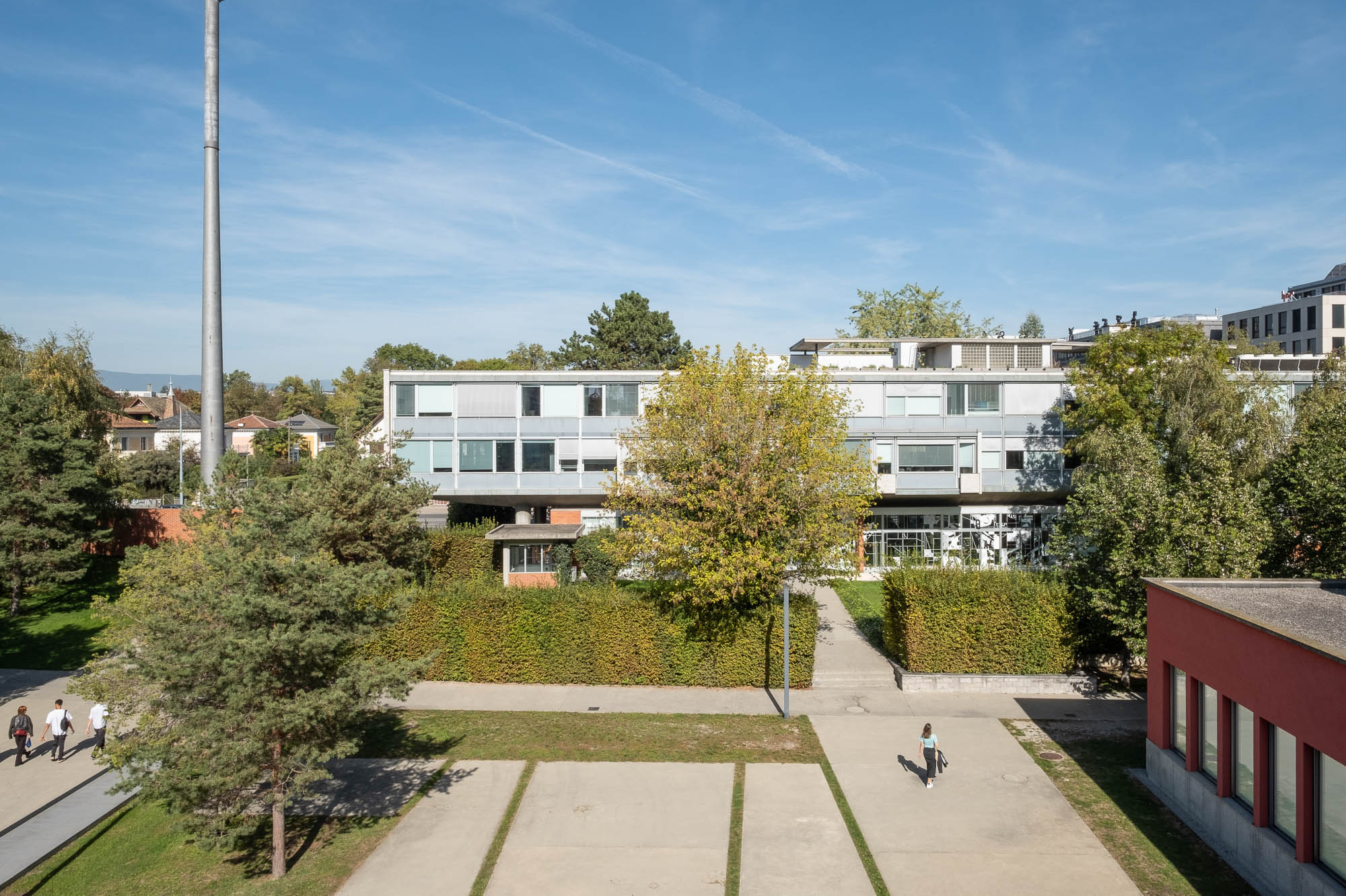 Exterior shot of a Modernist office block. In the foreground is a tree-lined yard in which people are walking.