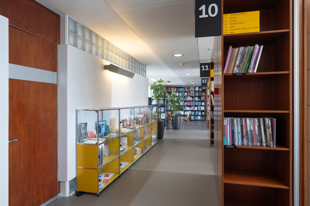 Interior of a library inside a former Modernist office building, with an interior wall to the left topped with glass bricks. At the back is a wall covered with magazine racks and plants. To the right are rows of wooden bookshelves.