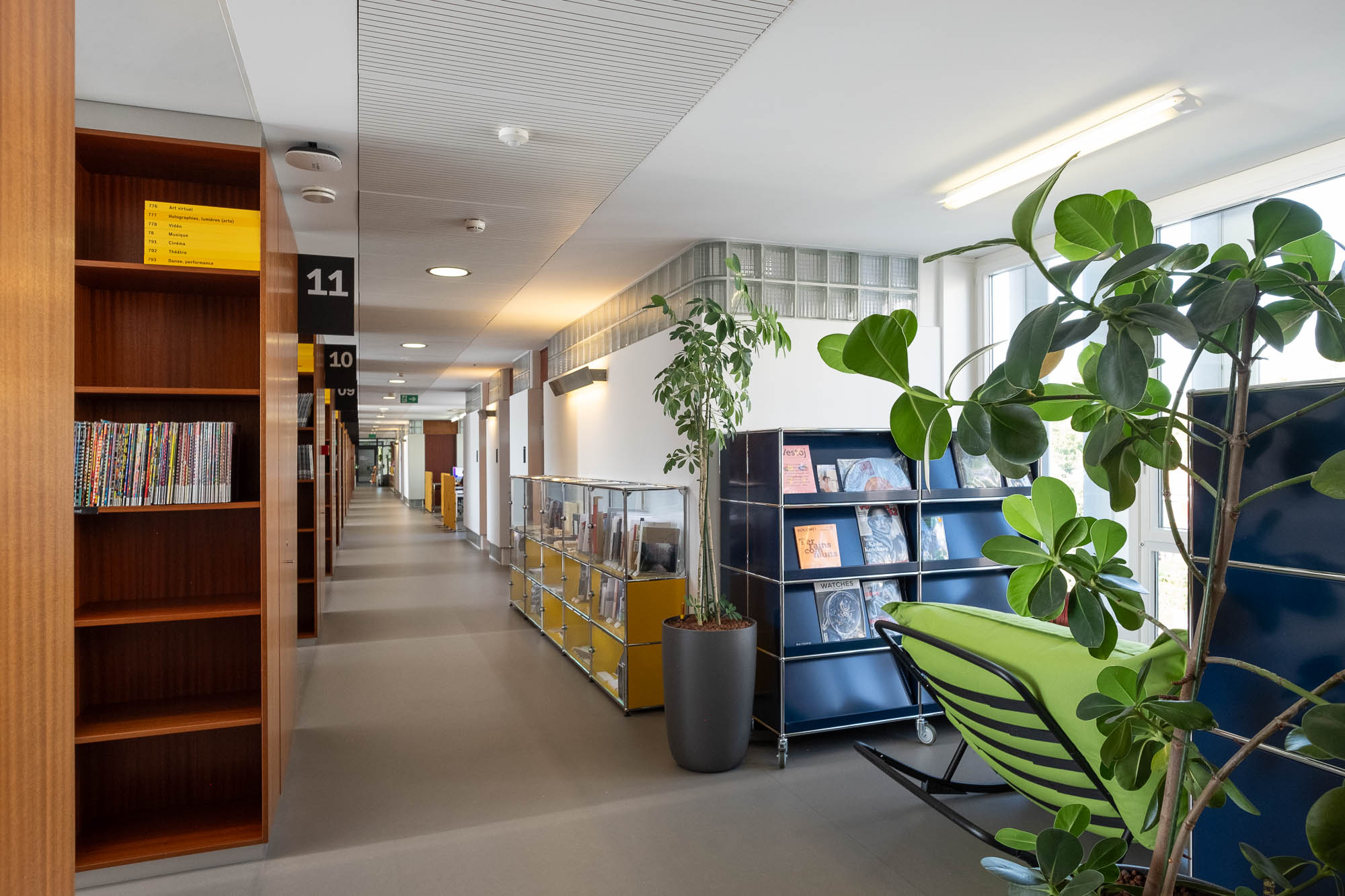 Interior of a library inside a former Modernist office building, showing an area with magazine racks on wheels, plants, comfy chairs and rows of bookshelves.
