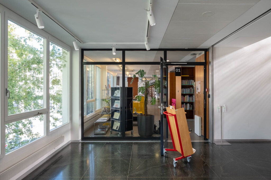 Interior of a library inside a former Modernist office building, showing the entrance to the library, a rectangular glass wall with a door. A book cart is set next to the door. Plants and bookshelves can be seen through the window.