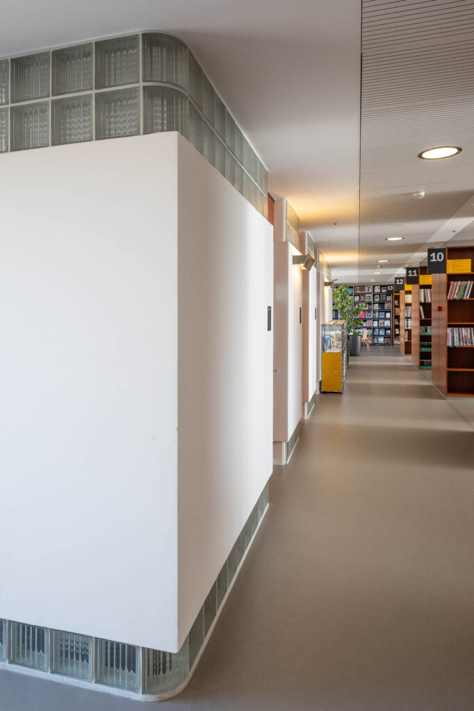 Interior of a library inside a former Modernist office building, showing the corner of an interior partition with glass bricks at the top and bottom. To the right are rows of bookshelves.