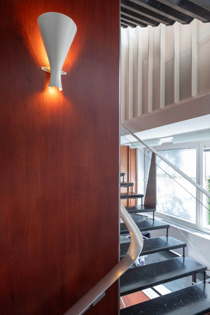 A staircase inside a Modernist office building, wrapping around a wood-clad central pillar on which is affixed a hourglass-shaped lamp.