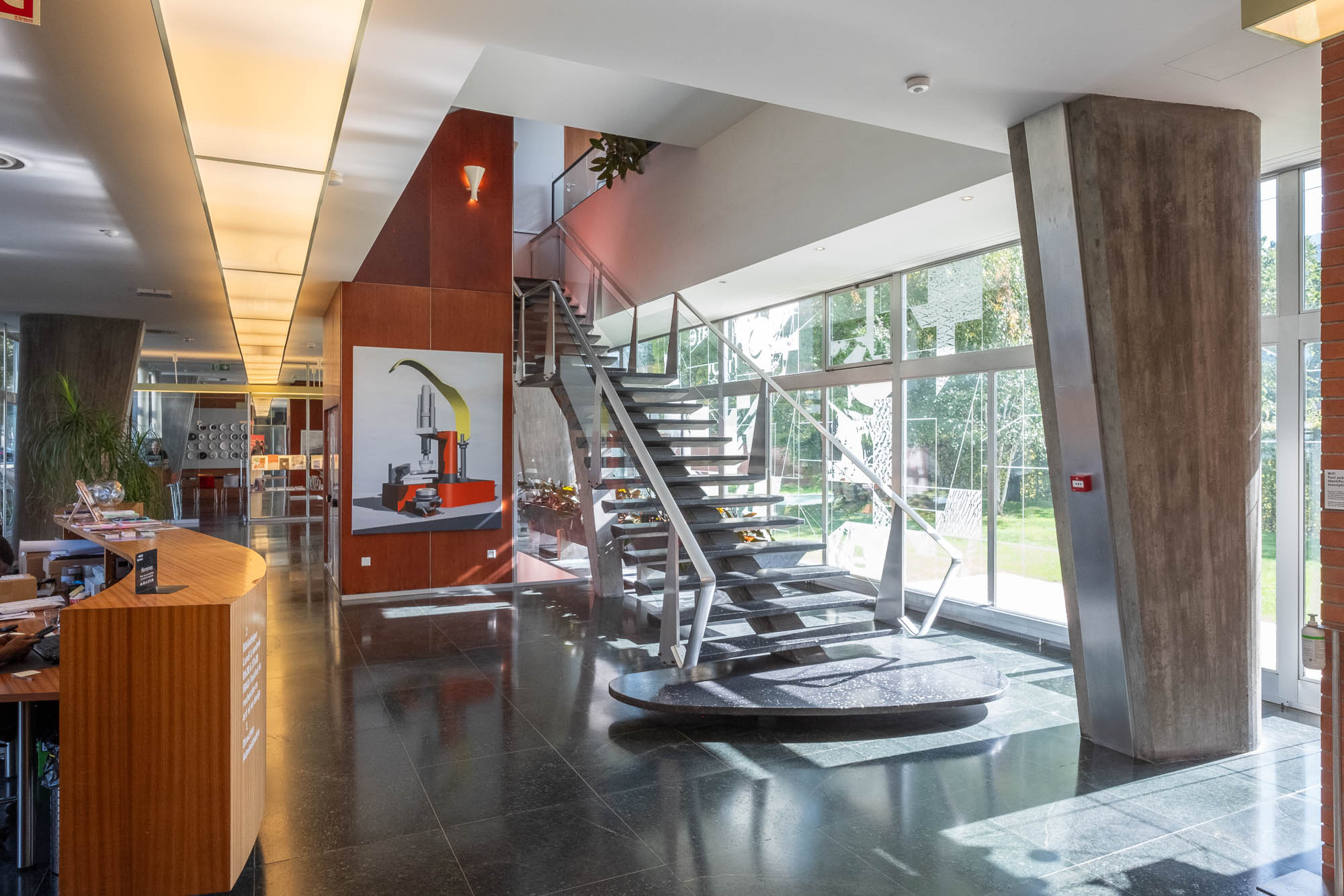 Interior of a Modernist office building reception area, showing a flight of stairs between tapered concrete columns, and windows at the back opening to a garden.