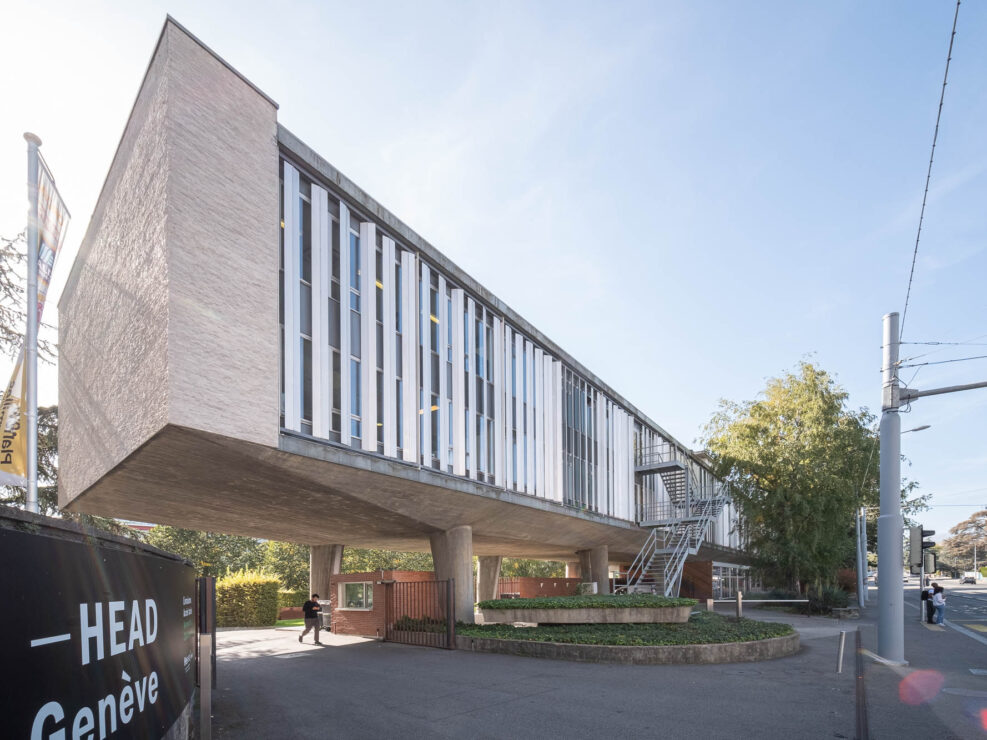 Wide-angle exterior shot of a Modernist office block on stilts. A person is walking underneath, next to a sign reading "HEAD Genève".