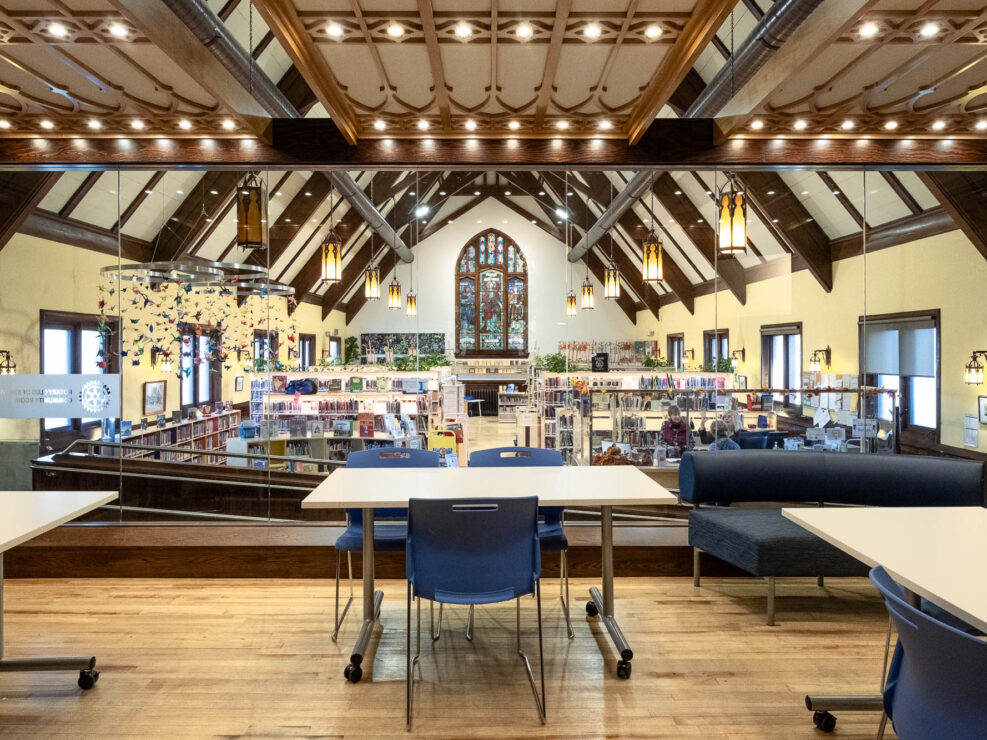 Interior of an English Neogothic church transformed into a library, viewed through a community room constructed in the former choir. The community room is separated from the nave by a glass partition, through which the library can be seen.