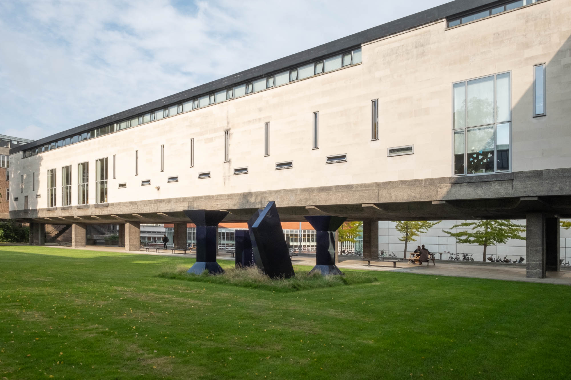 Exterior of a modernist university building, a three-storey structure on concrete stilts with a grass courtyard in the foreground.