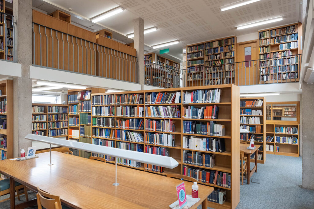 Interior of a modern university library with rows of wooden shelves surrounded by a mezzanine on which more shelves can be seen.
