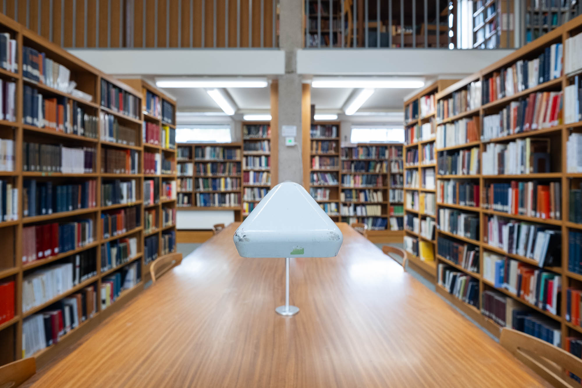 Out of focus image of a library interior with bookshelves on all sides and a table in the middle. The table is topped by a work lamp, the end of which is in focus.