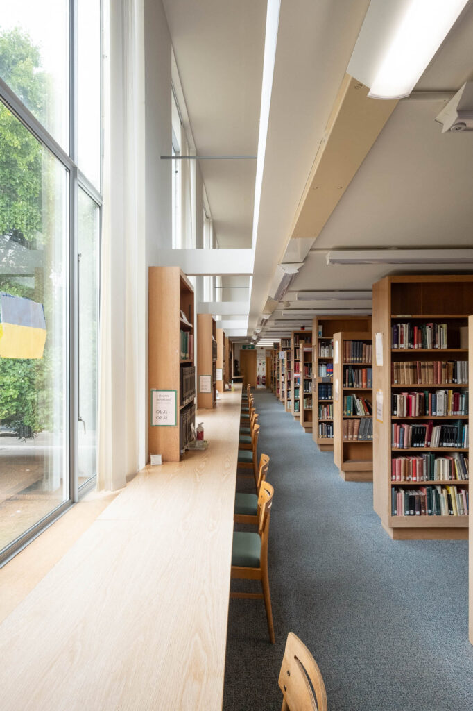 Interior of a modern university library, with a long table facing a row of windows. To the right are rows of bookshelves.