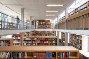 Interior of a modern university library with rows of wooden shelves surrounded by a mezzanine on which more shelves can be seen. To the left is the blurry figure of a person in motion.