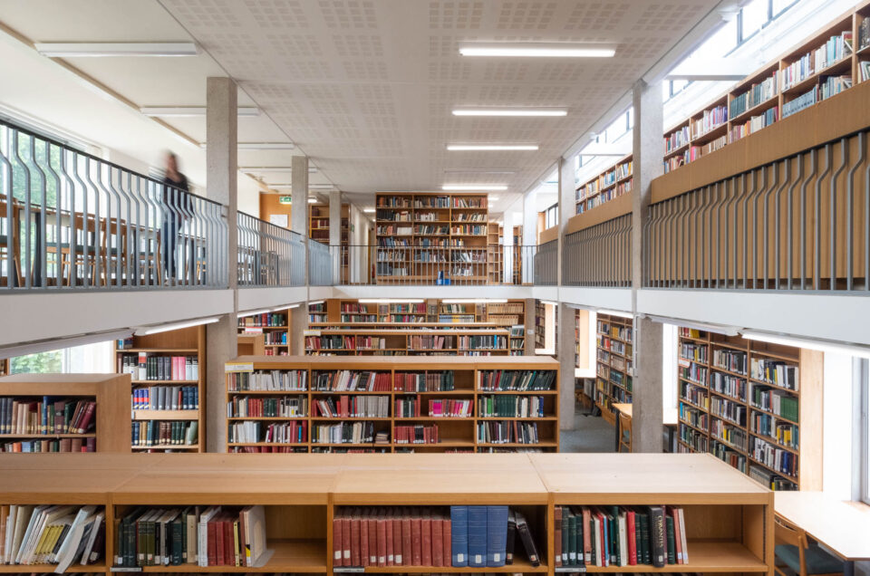 Interior of a modern university library with rows of wooden shelves surrounded by a mezzanine on which more shelves can be seen. To the left is the blurry figure of a person in motion.