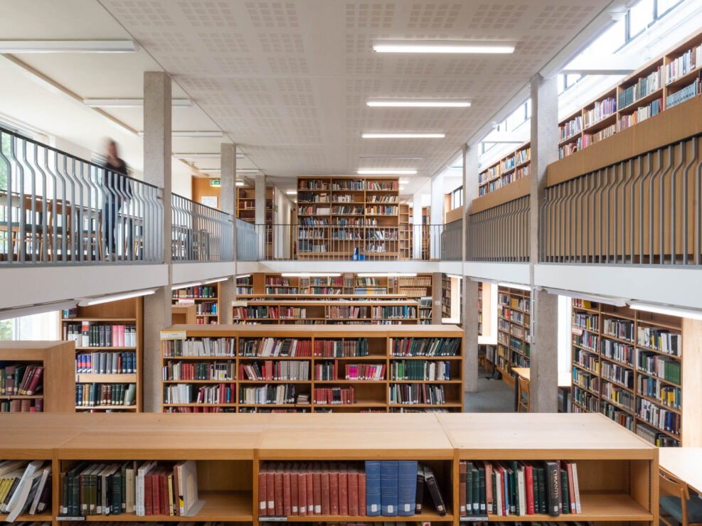 Interior of a modern university library with rows of wooden shelves surrounded by a mezzanine on which more shelves can be seen. To the left is the blurry figure of a person in motion.