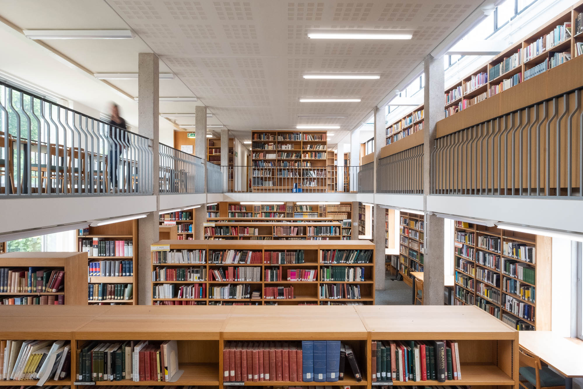 Interior of a modern university library with rows of wooden shelves surrounded by a mezzanine on which more shelves can be seen. To the left is the blurry figure of a person in motion.