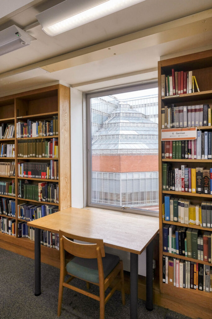 Interior of a library with a small work table facing a window and surrounded by bookshelves. Through the window, a pyramidal building of glass and brick can be seen.