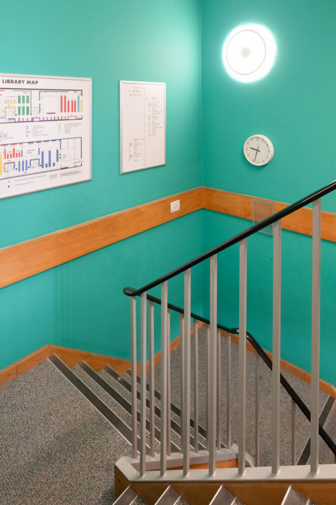 Interior of a staircase with walls painted a bright teal colour. On the walls are posters labelled "library map", a clock and a lamp.