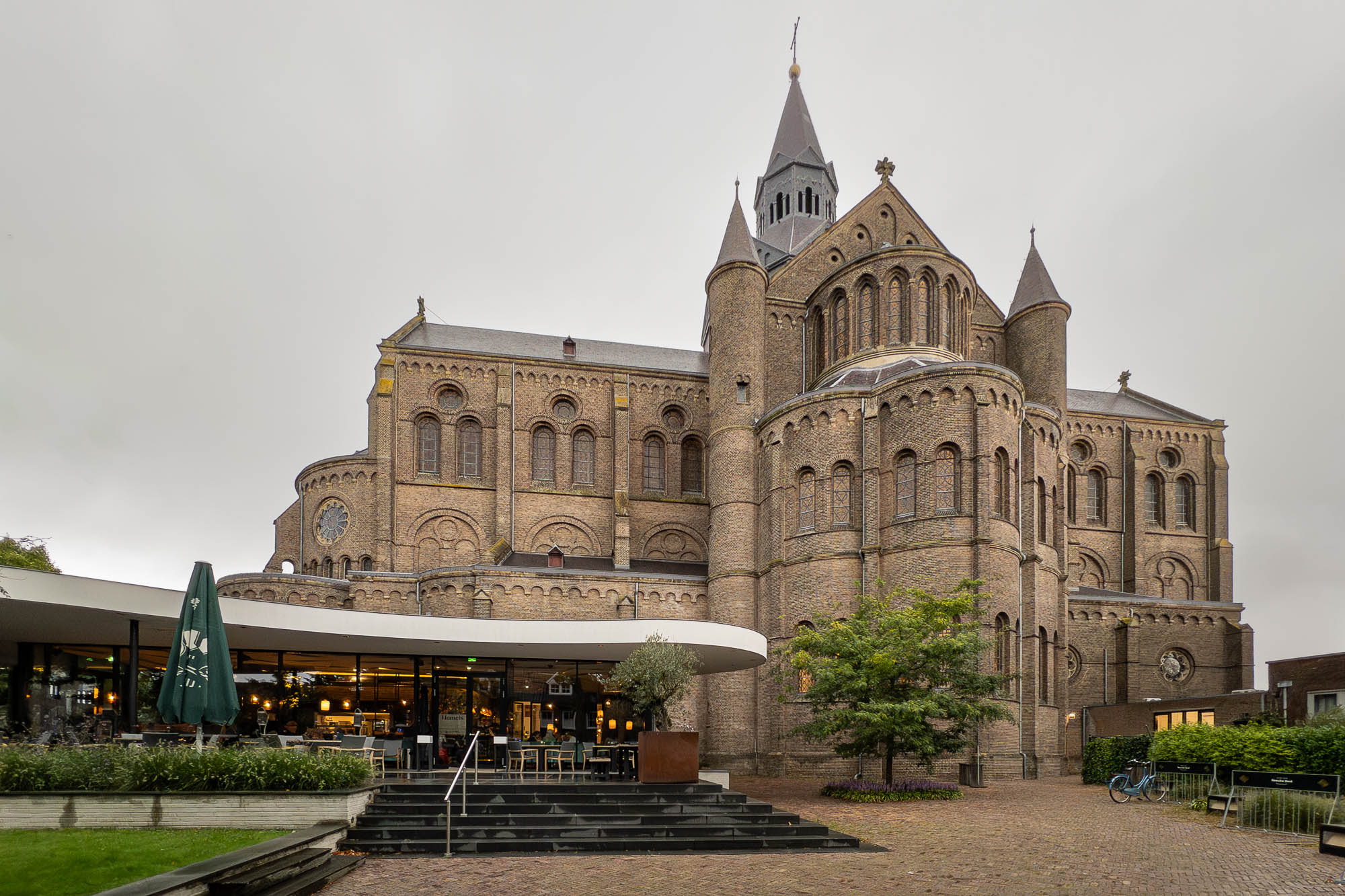 Exterior of a brick Neo-Romanesque church on an overcast day. A café under a curving roof stands in the foreground.