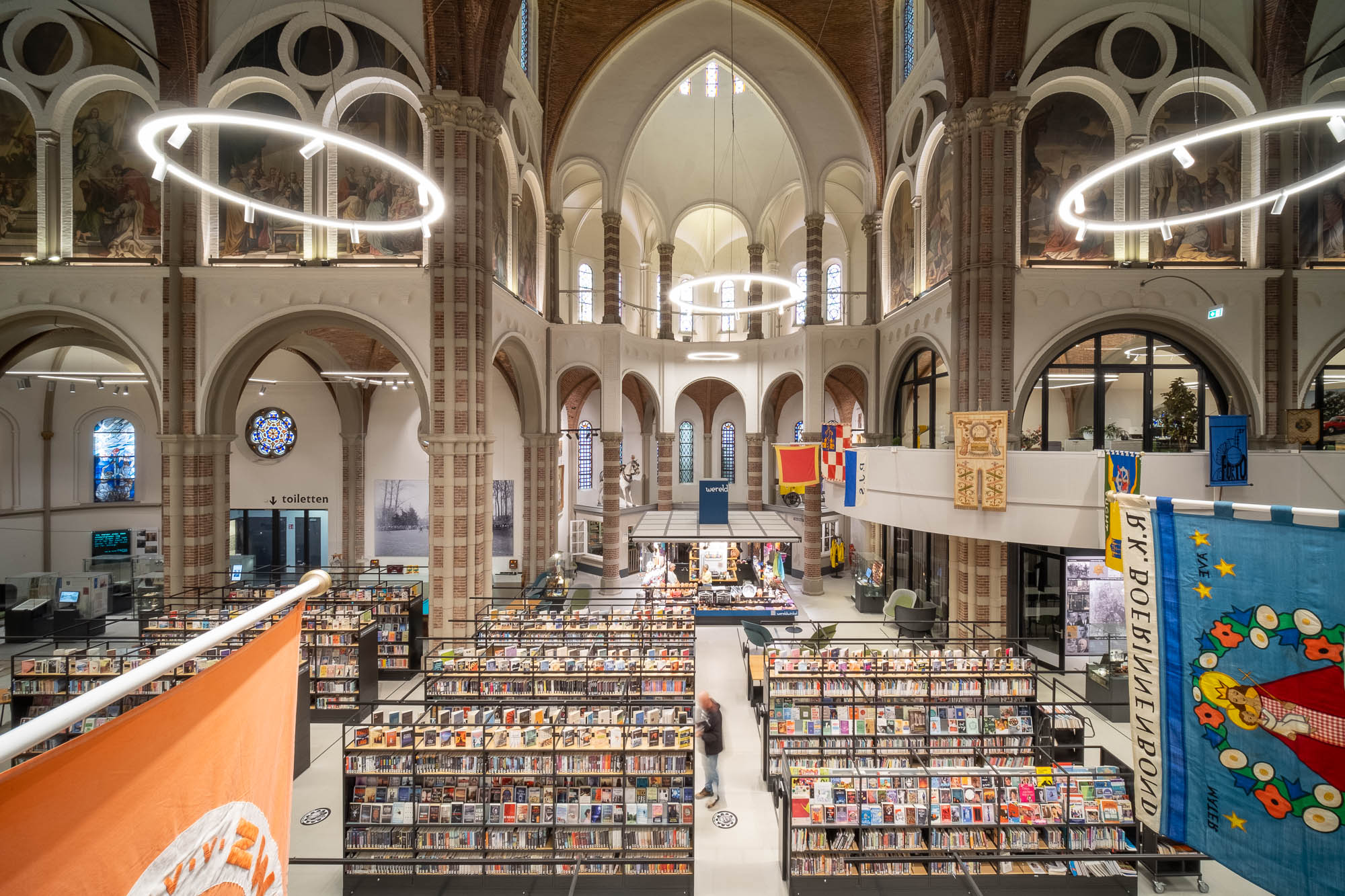 Interior of a brick church converted to a library with rows of contemporary bookshelves contrasting with the church interior. Flags are hanging from a curved mezzanine wrapping around the space.