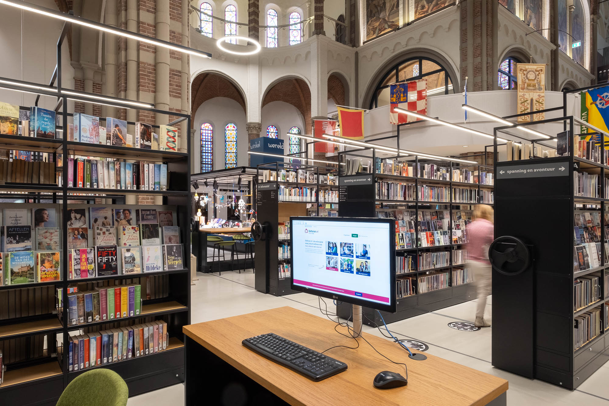 Interior of a brick church converted to a library, with rows of modern shelving on rails. A computer workstation sits in the foreground. A patron is moving between two rows of shelves, blurred by motion.