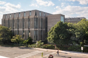 Exterior of a Modernist library, a rectangular building with glass facades covered by offset concrete lattice panels, surrounded by trees. A person walks on the street in front of the building