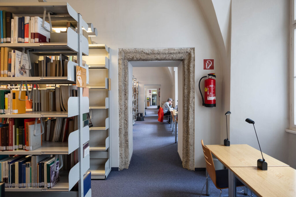 Interior of a library inside a Renaissance palazzo looking down an enfilade of doors. To the left are rows of metal bookshelves, to the right are narrow work tables, where a person works.