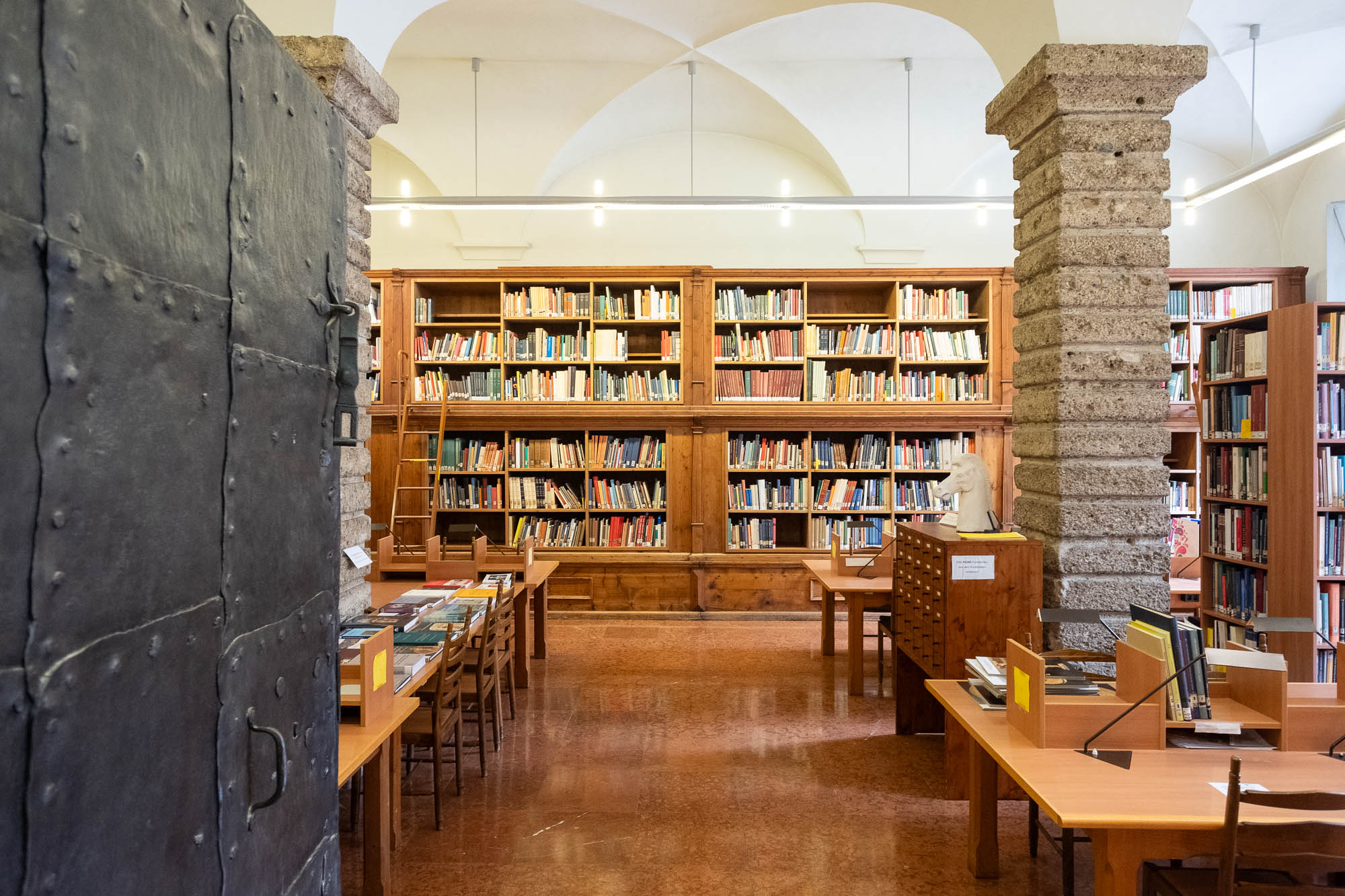Interior of a library inside a Renaissance palazzo, a vaulted room with high bookshelves and heavy rusticated stone columns. In the foreground is a heavy iron door.