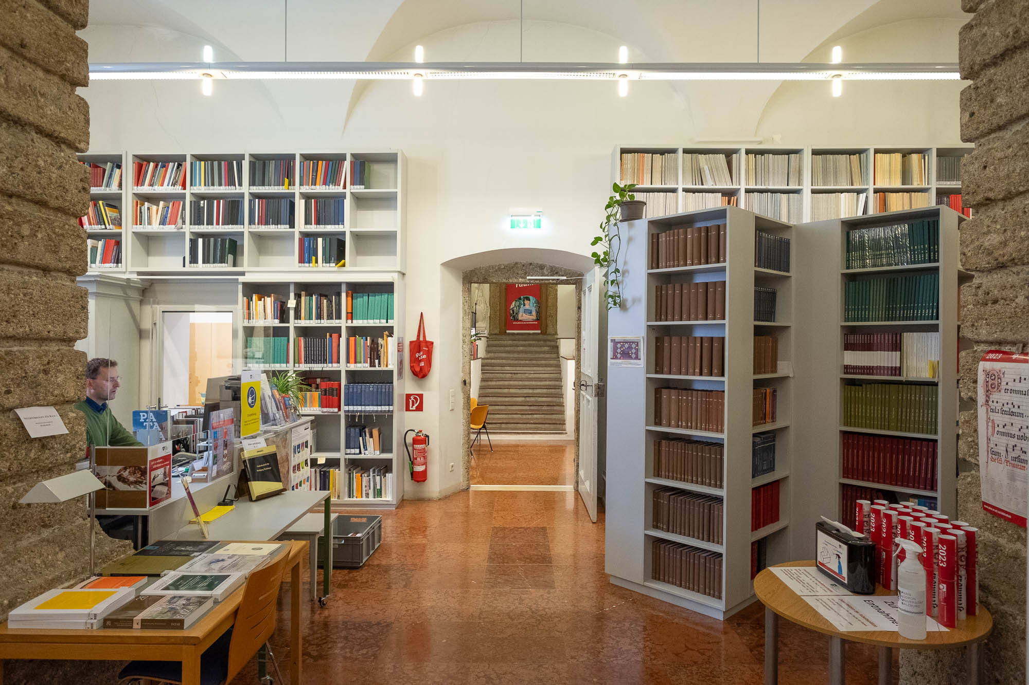 Interior of a library inside a Renaissance palazzo, a vaulted room with high bookshelves and heavy rusticated stone columns. To the side, a man works at a service counter.
