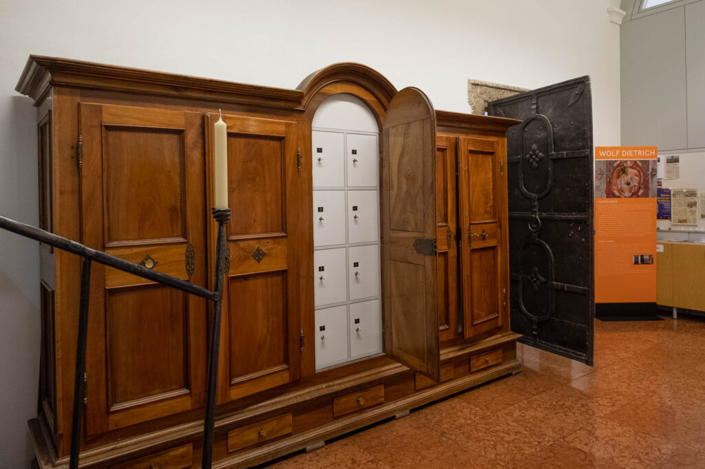 An anteroom to a library in a Renaissance palazzo, with a wooden wardrobe converted to hold metallic lockers. To the right is an open heavy iron door. In the front, a single candle is set in a stand built in to a stair railing.