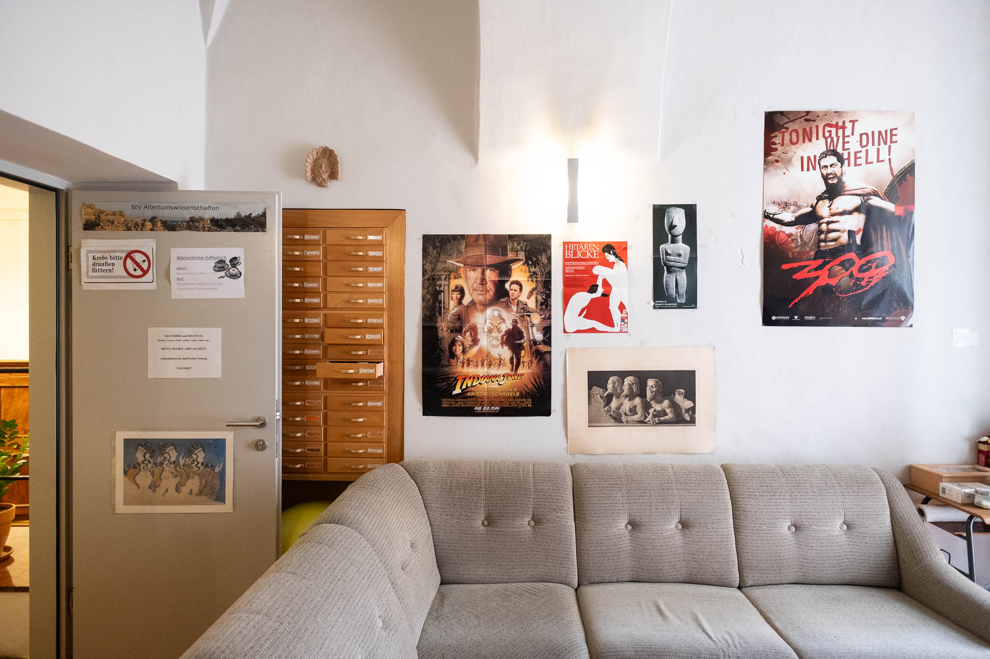 A recreation room with a couch and a wooden chest of drawers. The wall is decorated with several posters: a movie poster for Indiana Jones and the Crystal Skull, one for "300" with the text "Tonight we dine in hell", and others with black and white images of statues.