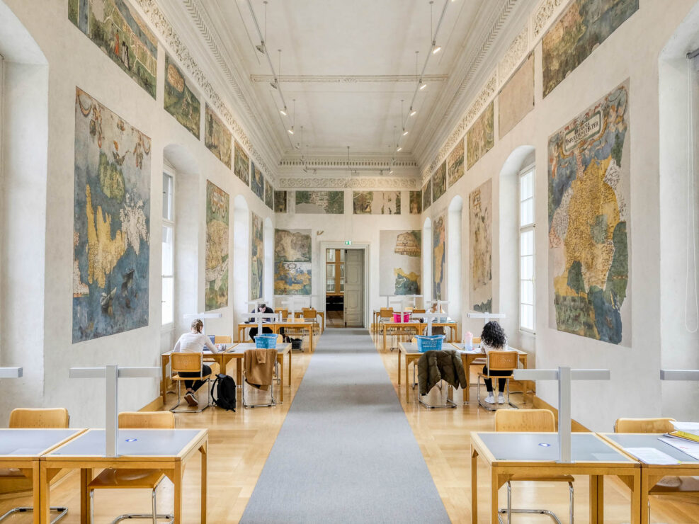 Interior of a library inside a Renaissance palazzo, showing a long and narrow reading room with work tables lining both walls. Between large vaulted windows, the walls are decorated with frescoes depicting maps.