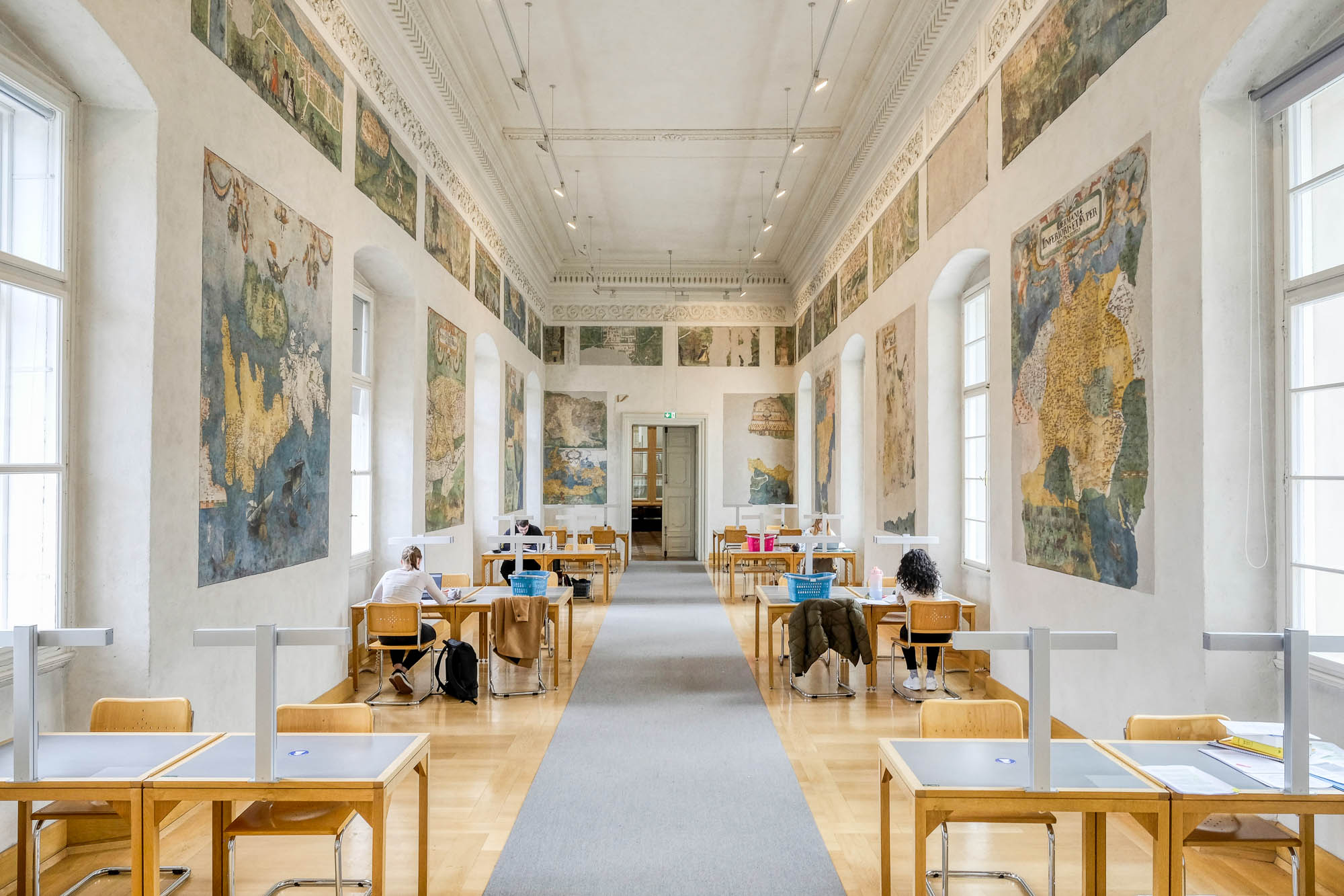 Interior of a library inside a Renaissance palazzo, showing a long and narrow reading room with work tables lining both walls. Between large vaulted windows, the walls are decorated with frescoes depicting maps.